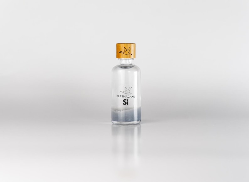 Puede incluir: Una botella de vidrio transparente con una tapa de madera. La botella tiene una etiqueta con el texto "PLASMAGAN: Si" y un logotipo de un p&aacute;jaro. La botella est&aacute; llena de un l&iacute;quido azul claro. La tapa de madera tiene un logotipo de p&aacute;jaro grabado.