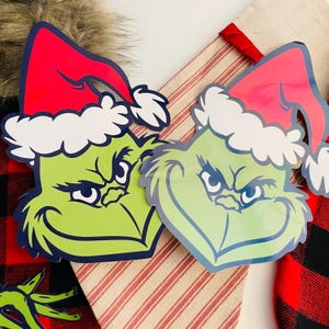 May include: Two green Grinch face cutouts wearing red Santa hats with white trim. The Grinch faces have a dark blue outline and are set against a red and white striped background and a red and black plaid background.