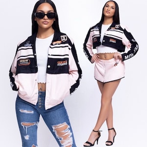 Puede incluir: Una chaqueta corta rosa y negra con varios parches y logotipos, incluyendo "GEESAS STUDIO" y "PERFECTION MOTION". La chaqueta tiene una cremallera y un cierre de botones en la parte delantera. La modelo lleva la chaqueta con una camiseta blanca y unos vaqueros rotos.
