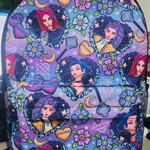 May include: A backpack with a repeating pattern of stylized female portraits in heart-shaped frames, flowers, and crescent moons. The color scheme includes purple, blue, and pink. The text "GOLDEN" and "AKE DOWN" are visible.