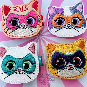 Könnte beinhalten: Vier Filz-Applikationen in Form von Katzenköpfen mit unterschiedlichem Fell und Masken. Die Katzen haben orange, lila, weißes und gelbes Fell. Die Masken sind rosa, orange, türkis und lila.