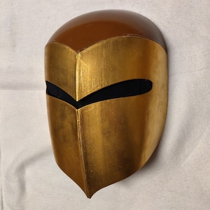 May include: A gold and brown helmet with a black visor. The helmet is made of a hard material and has a smooth finish.