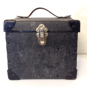 May include: A vintage black box with a metal clasp and corner protectors. The box has a brown leather handle and is likely used for storage or carrying items. The box is square-shaped and shows signs of wear.