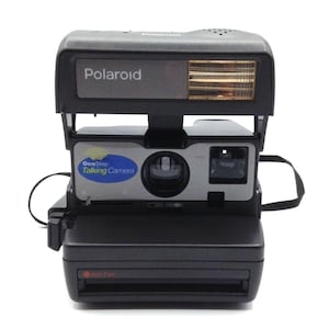 Peut inclure: Appareil photo Polaroid OneStep Talking Camera noir avec flash intégré. Le devant est argenté et il y a une sangle noire. Le texte "Polaroid" est visible. Il utilise du film 600.