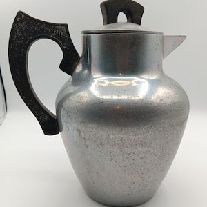 May include: Vintage silver-toned metal coffee pot with a dark brown handle and lid grip. The pot has a rounded body, a spout, and a lid. The handle is curved and attached to the side of the pot. The lid has a small, rectangular grip.