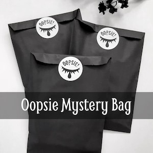 May include: Three black paper bags with white "Oopsie!" stickers. The bags are stacked on top of each other. The text "Oopsie Mystery Bag" is printed in white on a black banner below the bags.