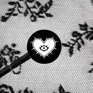 May include: Black and white sticker with a heart shape with a single eye in the center. The heart has a white outline and is surrounded by spikes. The sticker is on a black background.