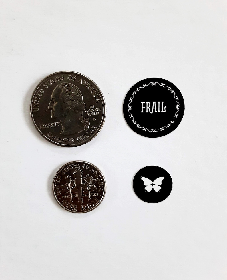 May include: Three black and white round stickers. The first sticker has a decorative border and the word "FRAIL" in white text. The second sticker has a butterfly design. The third sticker has a design of a tree with the words "UNITED STATES OF AMERICA" and "ONE DOLLAR" around the tree.