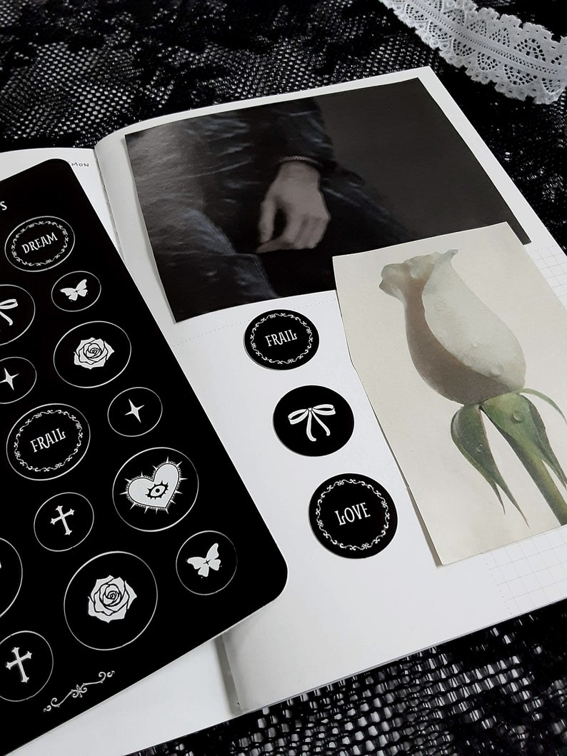 May include: A black planner with white stickers featuring words like "Dream", "Frail", "Love", and "Cross". The stickers are arranged on a white page with a black and white photo and a white flower illustration.
