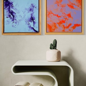 May include: Two square abstract paintings in wooden frames. One features purple swirls on a light blue background, the other has orange patterns on a lavender backdrop. A white, curved side table holds a small cactus in a pink pot and two open books.