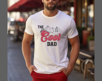 May include: A white short-sleeved t-shirt featuring a black line-art illustration of mountain peaks and pine trees above the text "THE" in black, "Cool" in a distressed red font, and "DAD" in a distressed black font on the chest.