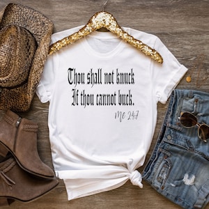 May include: White t-shirt with a black graphic that reads "Thou shall not knock if thou cannot buck. Mc 24:7". The shirt is laid out on a wooden surface with a straw hat, brown boots, and denim shorts.