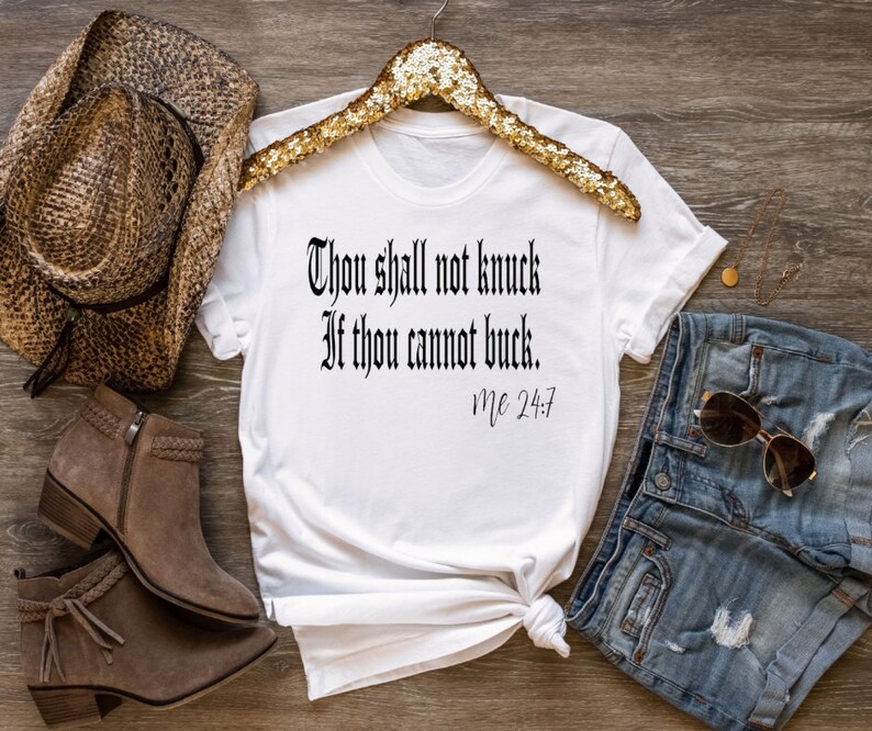 May include: White t-shirt with a black graphic that reads "Thou shall not knock if thou cannot buck. Mc 24:7". The shirt is laid out on a wooden surface with a straw hat, brown boots, and denim shorts.