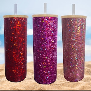 May include: Three glitter-covered tumblers with bamboo lids and clear straws. The tumblers are cylindrical and feature vibrant glitter in shades of red, purple, and a reddish-brown. The background shows a beach scene with sand and a blurred ocean.