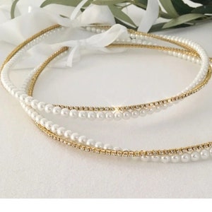 May include: Two gold and white pearl wedding crowns. The crowns are made of a thin gold wire with small white pearls strung along the wire. The crowns are intertwined.