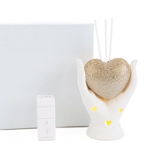 May include: White ceramic hands holding a gold heart-shaped diffuser with sticks. The diffuser is in a white box with a small white box with a tulip design.