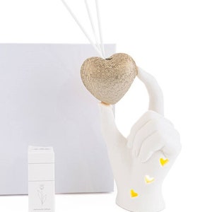 Könnte beinhalten: Weiße Keramikhand, die einen goldenen herzförmigen Diffusor mit drei weißen Schilfrohren hält. Die Hand hat drei gelbe Herzen ausgeschnitten.