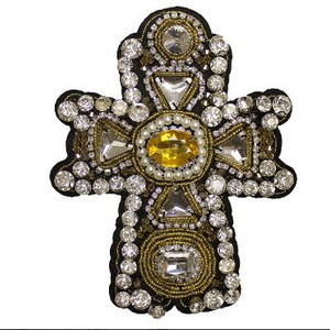 Può includere: Un ornamento a forma di croce in oro e argento con una grande gemma gialla al centro. La croce è ricoperta di strass e perline.
