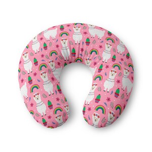 May include: A U-shaped nursing pillow featuring a repeating pattern on a pink background. The design includes white llamas with floral crowns, colourful rainbows, green cacti in pots, and small pink flowers scattered across the fabric surface.