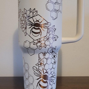 Könnte beinhalten: Ein weißer Edelstahlbecher mit einem schwarzen und goldenen Bienen- und Wabenmuster. Der Becher hat einen Deckel und einen Griff.