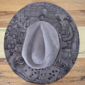 May include: A grey felt hat featuring a detailed black and white illustration on the brim. The graphic depicts a western saloon scene with cowboys, playing cards, revolvers, and cacti. The crown of the hat is plain grey with a side tassel detail.