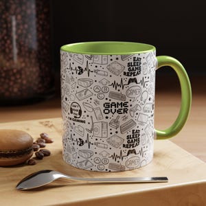 May include: A white ceramic mug with a green rim and handle. The mug features a black and white design of gaming icons and the text "Game Over" and "Eat Sleep Game Repeat".
