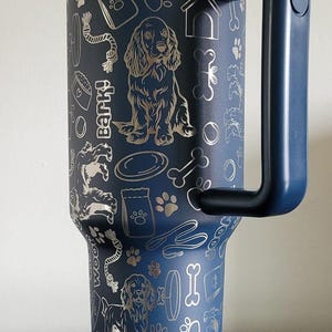 Puede incluir: Una taza de viaje de acero inoxidable azul con un asa negra. La taza está decorada con un diseño plateado de un perro, huesos, huellas de patas y otros artículos relacionados con los perros. El texto "Bark!" también está incluido en el diseño.