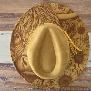 May include: A tan felt hat featuring a wide brim with a detailed brown print of a Highland cow and sunflowers. The crown is indented and encircled by a matching tan braided cord with dangling tassels on the right side of the hat's brim.