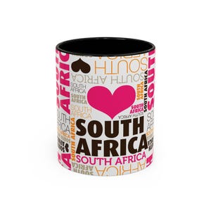 Könnte beinhalten: Weiße Keramiktasse mit schwarzem Rand und Henkel. Die Tasse zeigt ein Design aus den Worten "Südafrika", die in Herzform wiederholt werden, mit einem rosa Herzen in der Mitte. Die Wörter sind in verschiedenen Schriftarten und Größen.