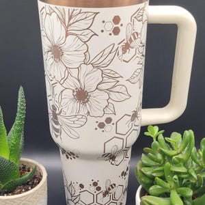Könnte beinhalten: Ein weißer Edelstahlbecher mit kupferfarbenem Deckel und Griff. Der Becher zeigt ein Blumenmuster mit Bienen und Wabenmustern in kupferfarbener Folie.
