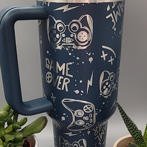 Könnte beinhalten: Eine blaue Reise-Thermoskanne aus Edelstahl mit einem weißen Deckel und einem schwarzen Griff. Die Kanne hat ein silbernes Gamer-Design mit Gamecontrollern, Blitzen und dem Schriftzug "Game Over".