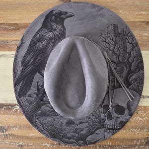 May include: A grey wide-brimmed hat featuring a detailed black graphic print of a raven perched on a branch, a human skull, and a desert landscape. The hat has a central crown indentation and a grey cord accent tied around the base.