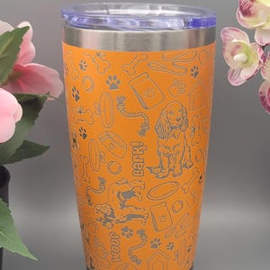 Puede incluir: Un vaso de acero inoxidable naranja con un diseño de temática canina. El vaso presenta una variedad de iconos relacionados con los perros, incluyendo huesos, huellas de patas, juguetes para perros y una caseta para perros. El texto "Bark" es visible en el vaso.