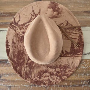 May include: A wide-brimmed tan hat featuring a dark brown printed illustration of a stag, a mountain range, and pine trees on the brim. The hat has a central crown indentation and a thin brown cord accent tied around the base of the crown.