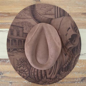 May include: A wide-brimmed brown felt hat featuring a detailed laser-engraved design on the brim. The illustration depicts a steer skull, a desert landscape with cacti, and a building with the word "TESKO" visible on the front facade.
