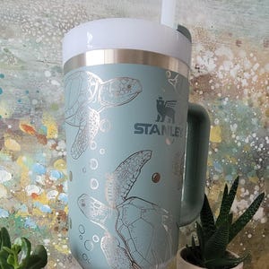 May include: A teal blue stainless steel travel mug with a handle and a white lid. The mug features a silver design of sea turtles and bubbles. The mug has the word "STANLEY" printed on the side.