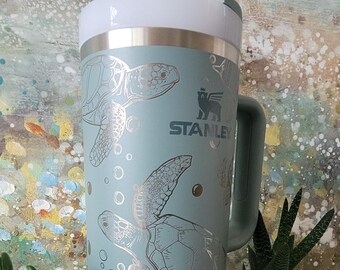 May include: A teal blue stainless steel travel mug with a handle and a white lid. The mug features a silver design of sea turtles and bubbles. The mug has the word "STANLEY" printed on the side.