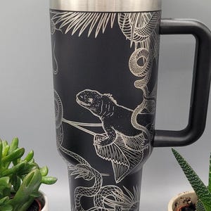 Op de afbeelding: Zwarte reistumbler van roestvrij staal met een zwarte handgreep. De tumbler heeft een wit ontwerp van een hagedis en slangen op een zwarte achtergrond.