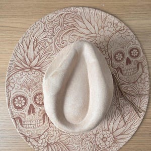May include: A beige felt hat with a wide brim featuring an intricate, dark brown laser-engraved pattern of sugar skulls, flowers, and swirling foliage. The crown is pinched, and a thin, light-coloured leather cord is tied around the base.