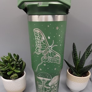 May include: A green stainless steel tumbler with a lid and handle. The tumbler features a laser-etched design of a moth, mushrooms, and stars.