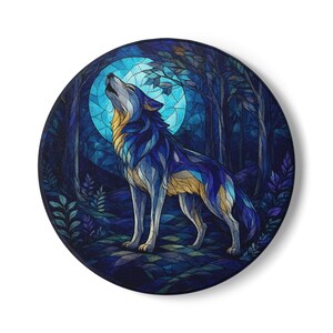 May include: A circular wall art piece featuring a stained glass style illustration of a wolf howling at a large, bright blue moon. The scene includes dark blue and purple forest trees and foliage, with the wolf rendered in shades of blue and gold.