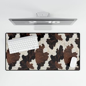 May include: A rectangular desk mat featuring a cowhide design in brown, black, and white. A white keyboard and mouse are on the mat. A computer monitor is positioned above the mat.