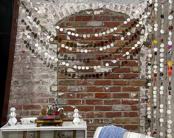 May include: A white bookshelf with two shelves, decorated with small white snowmen, framed pictures, and pink and red cards. A blue throw blanket is draped over a white and gray striped armchair. A brick wall with a whitewashed archway is decorated with a silver garland of circles.