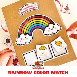 May include: A brown cardboard activity board featuring a colorful rainbow graphic with smiling clouds. The board includes the text RAINBOW COLOR MATCH and MadyBug. Several square tiles with illustrations like a strawberry, ladybug, and chick are present.