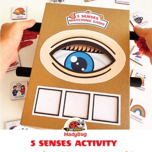 Puede incluir: Un tablero de juego 5 Senses Matching Game de cartón marrón con una gran ilustración de un ojo azul, una etiqueta que dice 5 Senses Matching Game y tres espacios rectangulares blancos vacíos, rodeado de pequeñas tarjetas cuadradas con iconos.