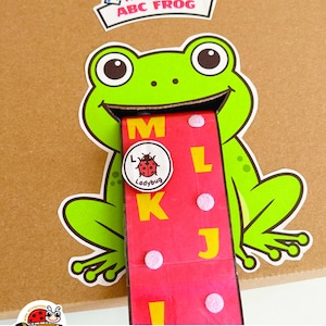 K&ouml;nnte beinhalten: Eine Papp-Aktivit&auml;t mit einem gr&uuml;nen Frosch mit weitem Maul und einer langen roten Zunge. Auf der Zunge befinden sich die gelben Buchstaben M, L, K, J, I und ein runder Marienk&auml;fer-Chip. Dar&uuml;ber steht The Hungry ABC FROG.