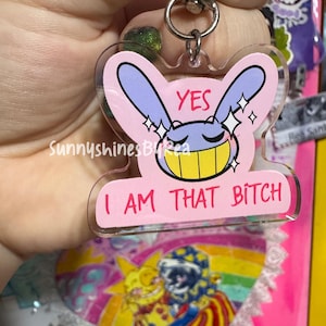 May include: A clear acrylic keychain featuring a pink background with a purple cartoon rabbit head that has a wide yellow grin and closed eyes. The text "YES" appears above the rabbit, and "I AM THAT BITCH" is printed in pink below it.
