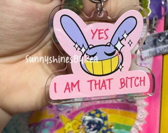 Op de afbeelding: Een doorzichtige acryl sleutelhanger met een roze achtergrond en een paarse cartoon konijnenkop met een brede gele grijns en gesloten ogen. De tekst "YES" staat erboven en "I AM THAT BITCH" is in het roze eronder gedrukt.