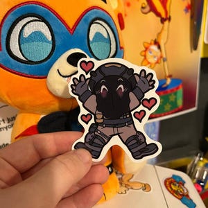 May include: A black and white sticker of a cartoon character wearing a helmet and a black and grey outfit. The character is holding out their hands and has red hearts around them.