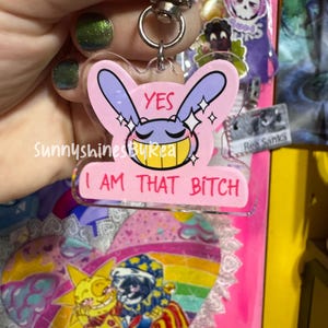 May include: A clear acrylic keychain featuring a cartoon character with purple ears and a yellow grin. The design includes the text YES and I AM THAT BITCH in pink and black lettering, accented with white sparkles on a light pink background.